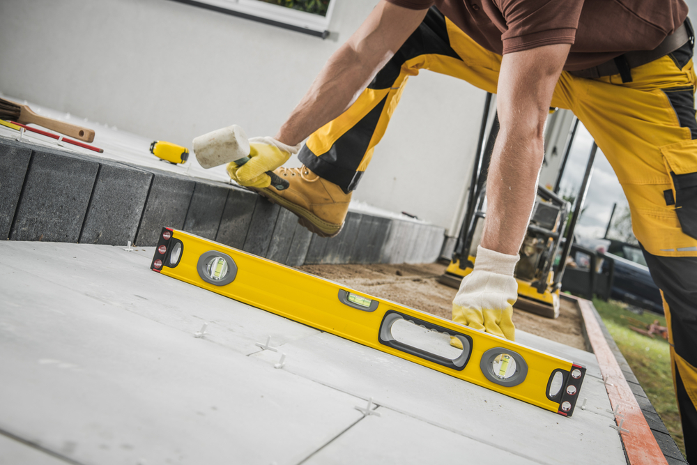 Contractor Laying Concrete Tiles During the Construction of a Terrace in the Backyard Garden of a Private Residential House and Checking Their Evenness with a Spirit Level Tool.